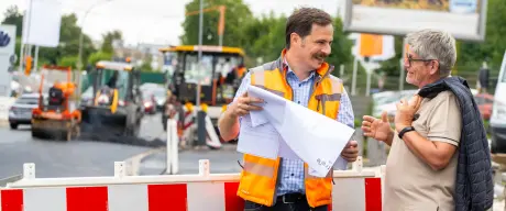 Mann mit Warnweste unterhält sich mit einem anderen Mann. Im Hintergrund steht eine Schrankenzaun und Baustellenfahrzeuge